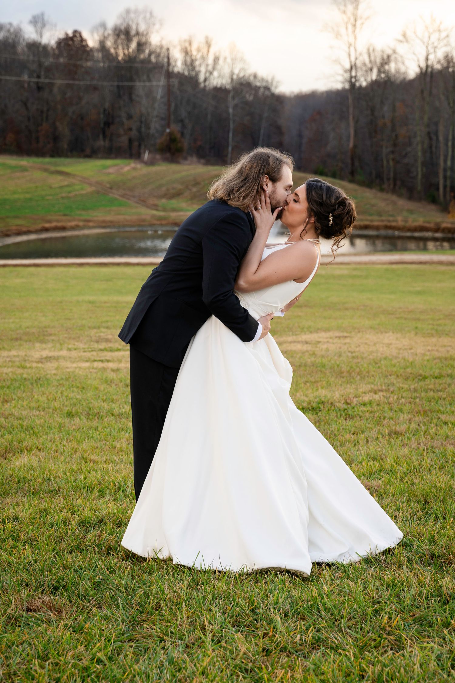 Groom dipping his bride for a kiss beside a pond in warm sunset light.