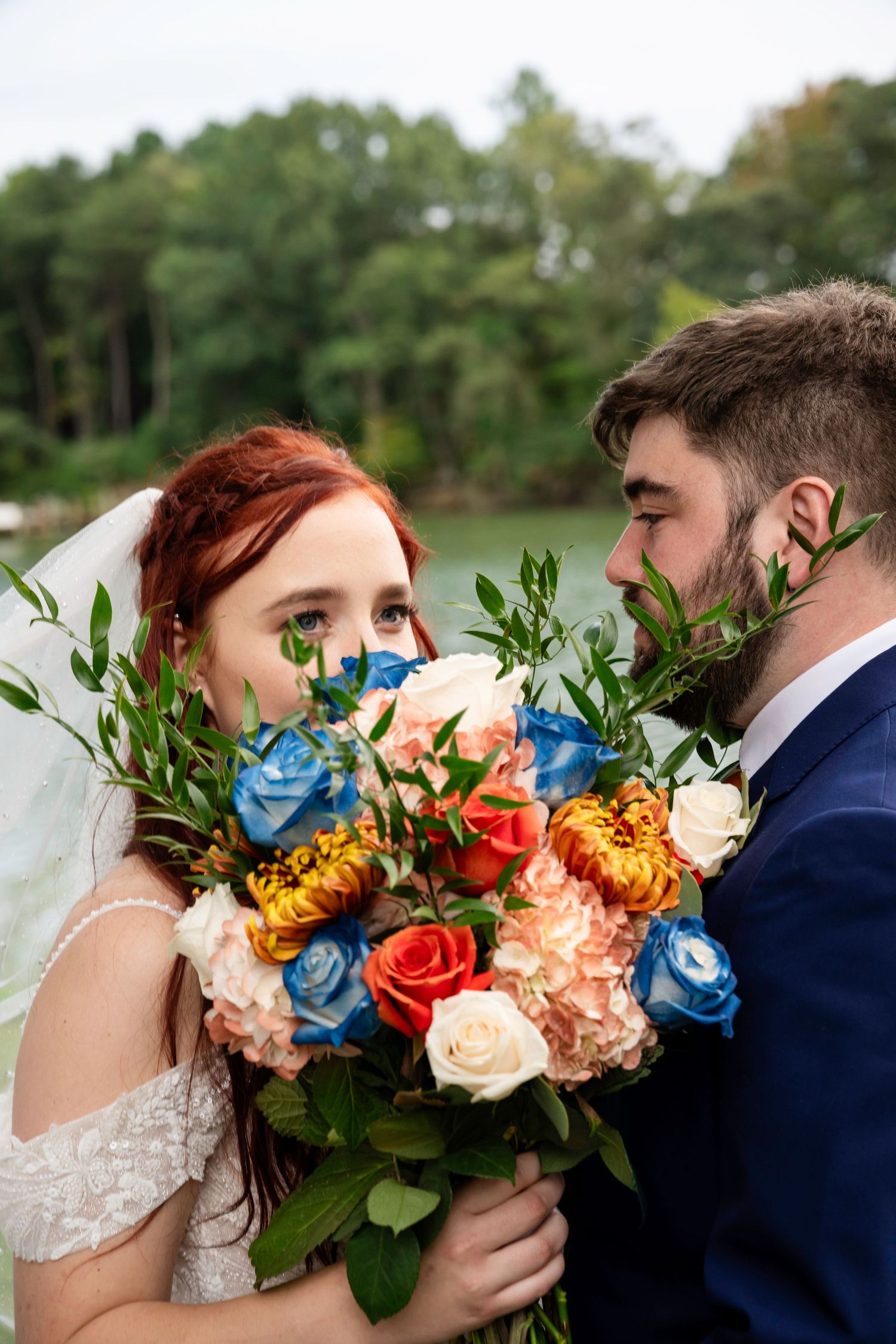 Bride and groom smiling over a colorful bouquet of blue, orange, and peach roses.