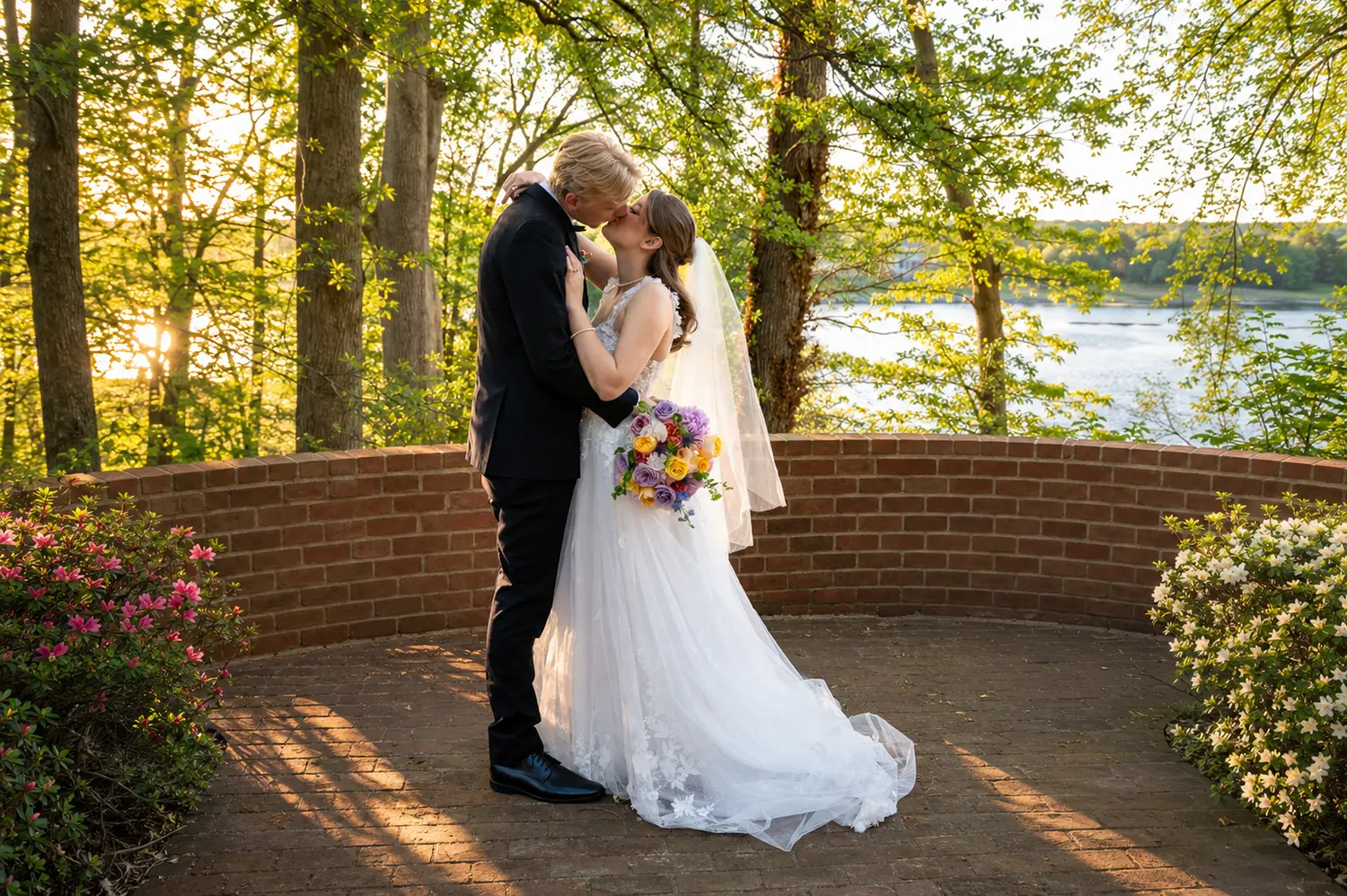 Bride and groom kissing on a brick terrace beside a peaceful lake at sunset.