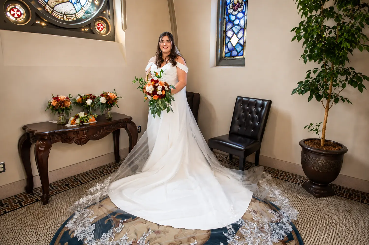 Bride holding a vibrant bouquet beside stained glass windows before her wedding ceremony.