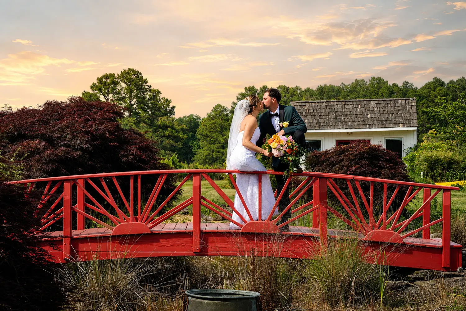 Bride and groom kissing on a red garden bridge at sunset with colorful flowers surrounding them.