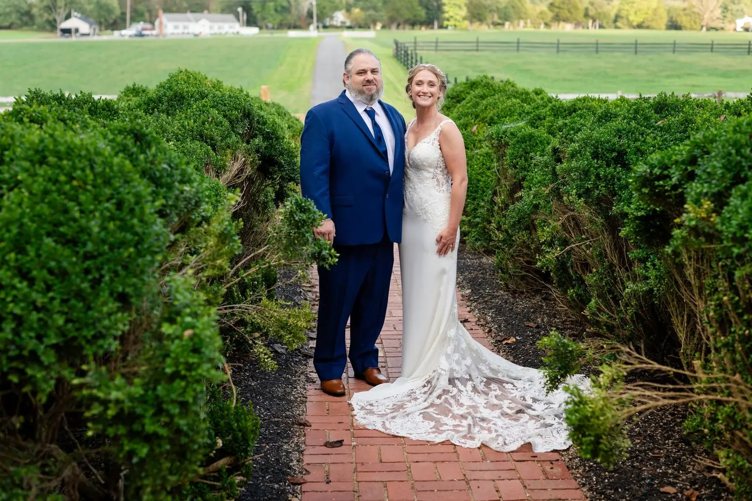 Bride and groom walking together along a brick garden path framed by tall green hedges.