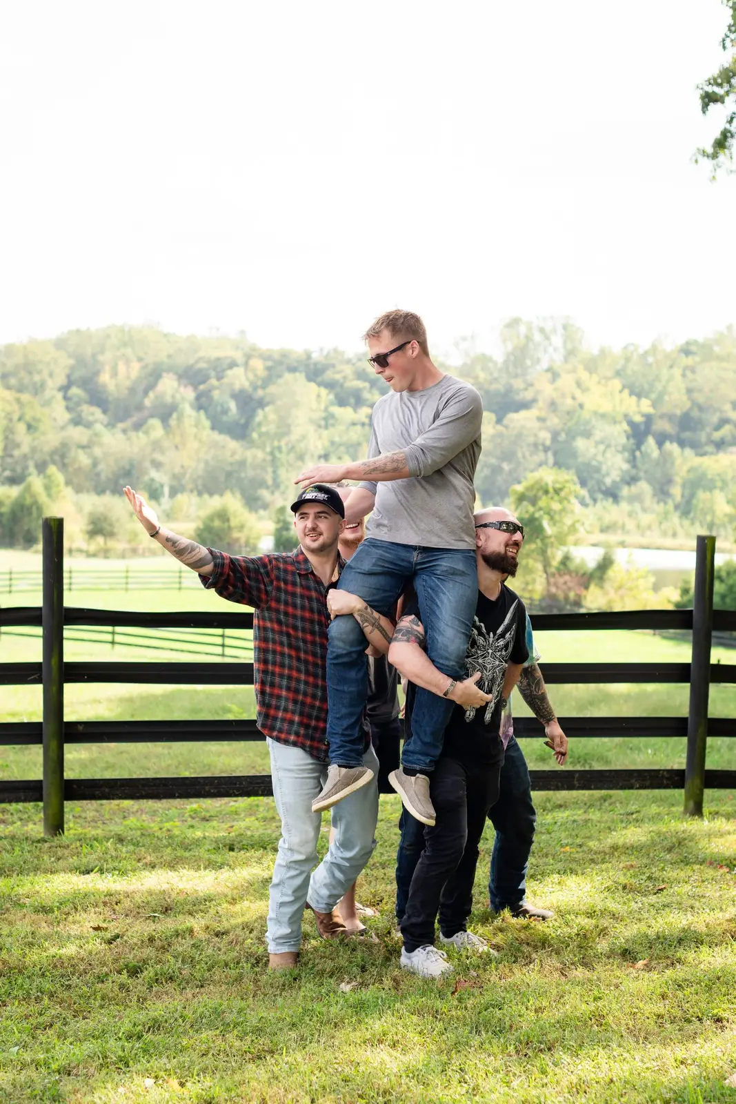 Four groomsmen laughing together beside a wooden fence in the sunny countryside.