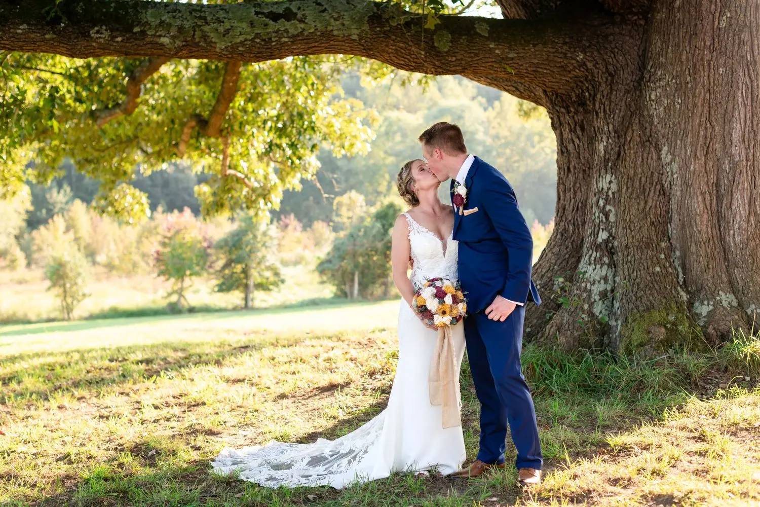 Bride and groom kissing beneath a large sunlit oak tree while the bride holds a colorful bouquet.
