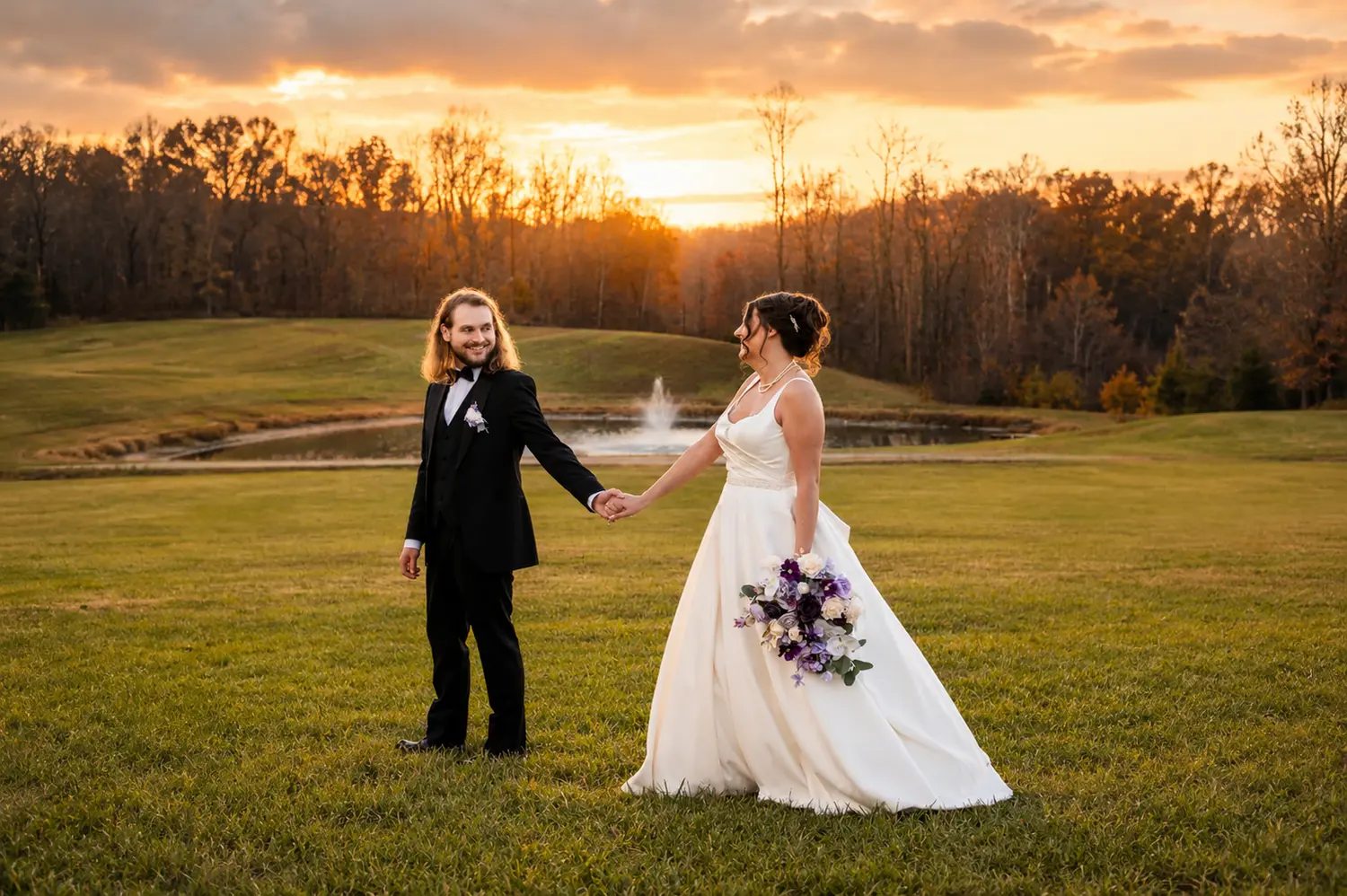 Bride and groom walking hand in hand beside a pond and fountain in warm sunset light.