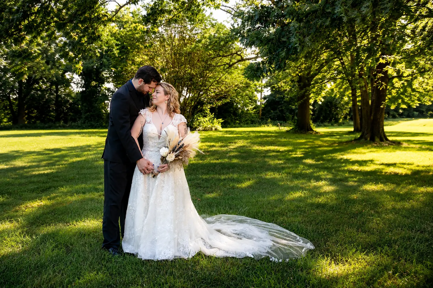 Newlyweds embracing on a wooded riverbank surrounded by lush summer greenery.