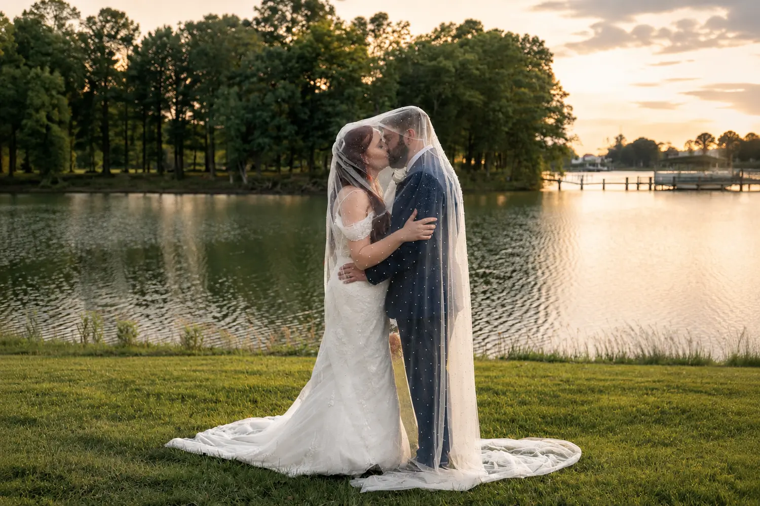 Bride and groom kissing beneath a pearl-detailed veil beside the river at sunset.