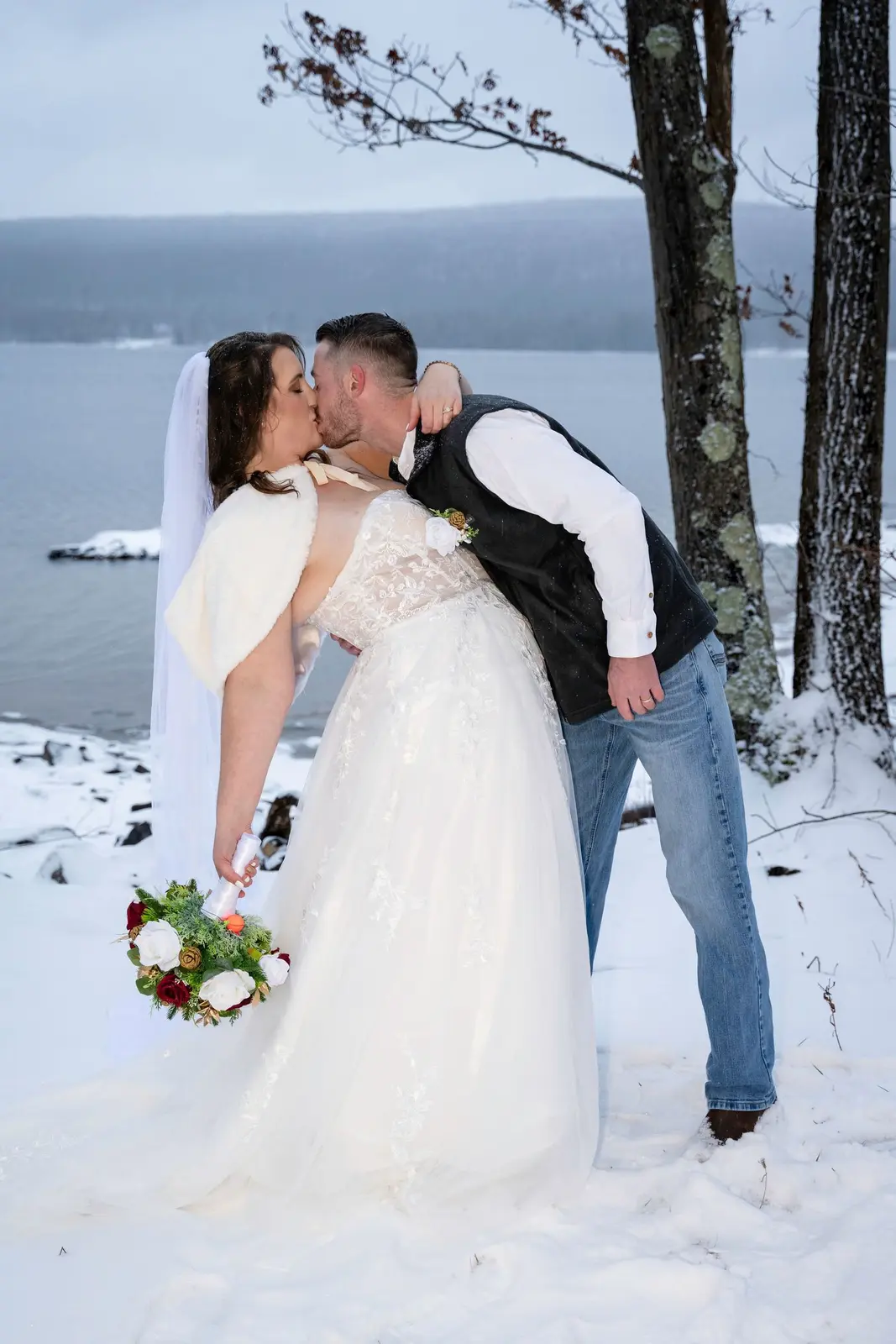 Bride and groom sharing a kiss beside a snowy lake while the bride holds a red and white bouquet.