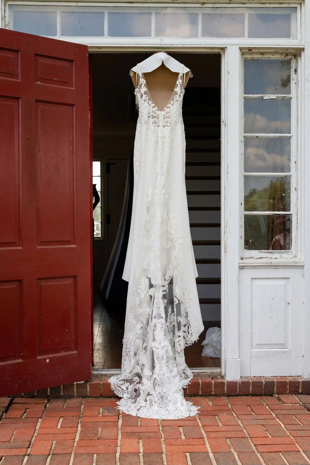 White lace wedding dress hanging between red and white doors on a brick porch.