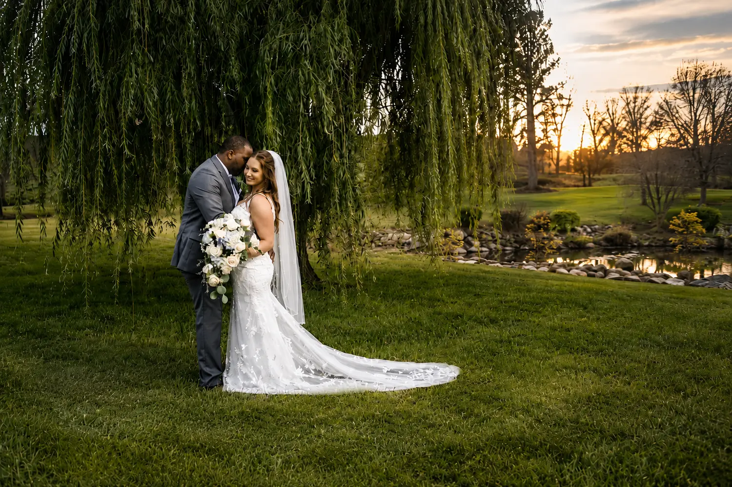 Bride and groom kissing beneath a sweeping weeping willow beside a quiet pond.