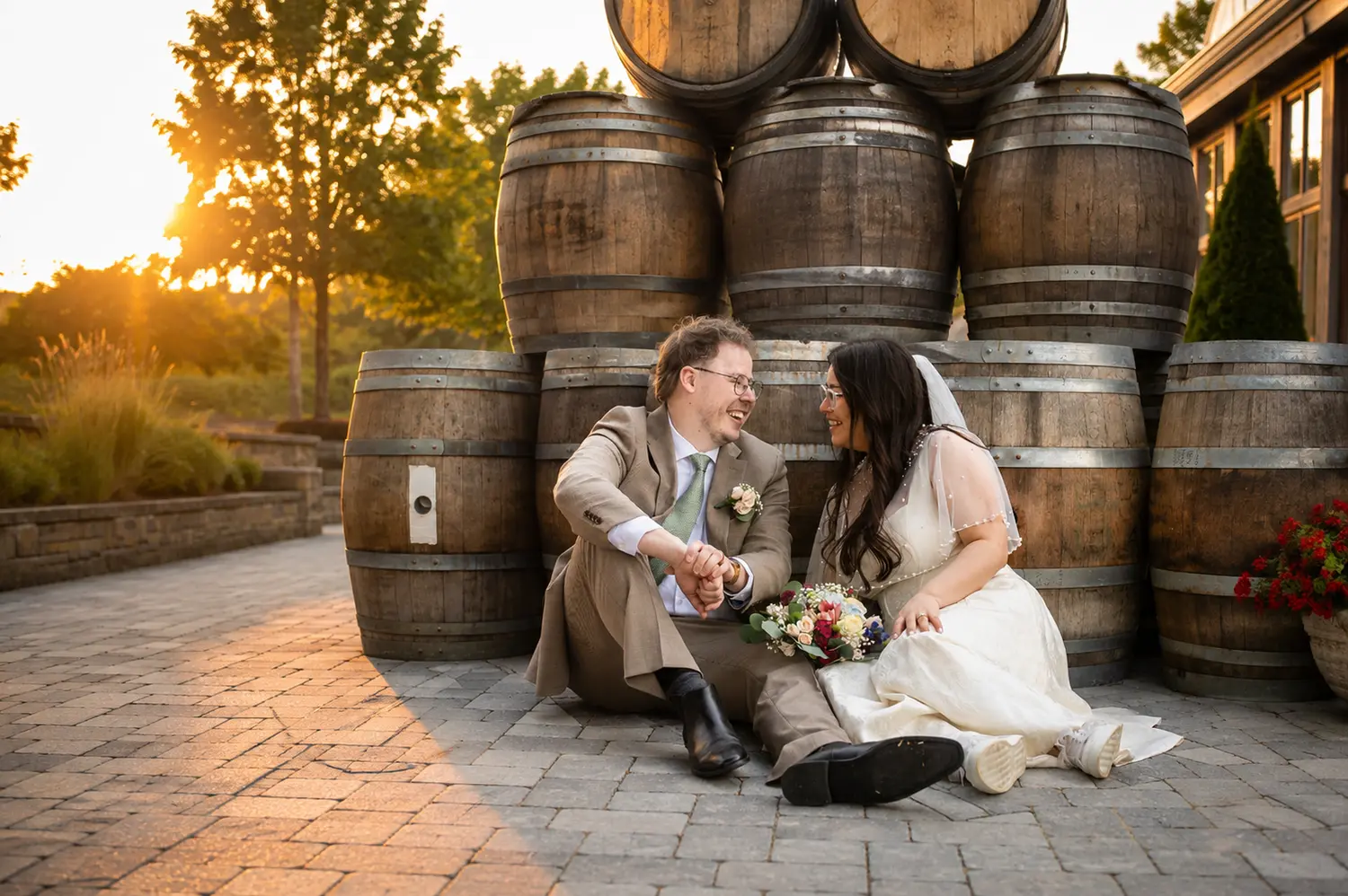 Bride and groom laughing together in front of rustic wine barrels at golden hour.